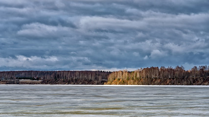 A spring landscape with a stormy sky over a frozen river and a lone ray of the setting sun
