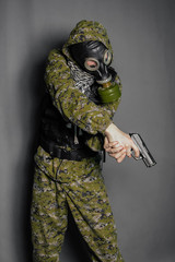 A soldier in camouflage uniform, body armor, in a gas mask with a hood on his head, holds a gun in his hands. Cosplay man, stalker. Studio photo on a gray background.