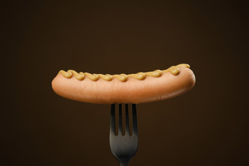 sausage with mustard on a black fork. dark brown background. isolated