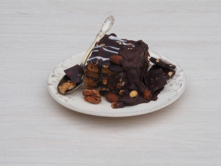 Chocolate cake with nuts and icing in a white plate with a spoon on a light background closeup. A piece of biscuit with pieces of chocolate and different nuts. Sweet pastry  with copy space