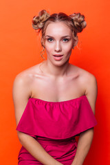 The girl in a red dress on a orange background in the studio. Blonde girl with two hair knots looking to the camera and smiling.