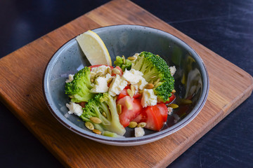 Two plates of vegetable salad with feta cheese, cucumbers, tomatoes, and broccoli served in bowl on rustic wooden board.