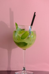 Mojito alcohol cocktail with plastic straw, tropical beverage with ice, mint and lime on pink background, copy space.