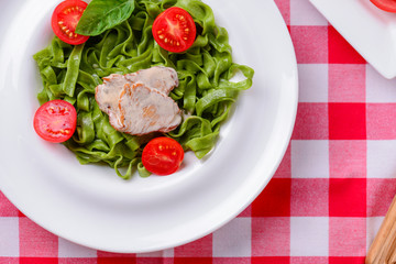 Tagliatelle pasta with pesto, tomatoes and meat stake served on white plate over red plaid tablecloth.