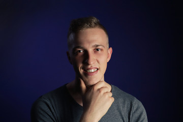 Obraz premium Young caucasian blonde smiling man in grey t shirt on dark purple background touching his chin