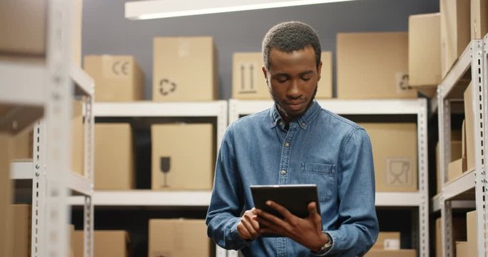 African American handsome young postman standing in postal store with parcels and tapping on tablet device. Male worker with computer doing registration in delivery room full of boxes.