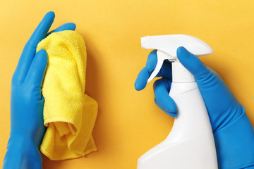 Hands in protective rubber gloves holding a spray bottle with detergent and a cloth on a yellow...