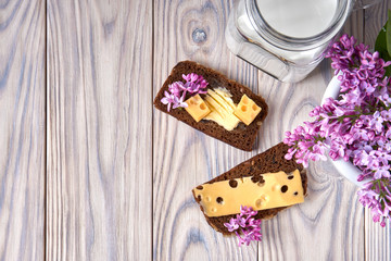 Dark multigrain bread made from rye, wheat, oats, barley with sunflower, flax seeds. Milk, cheese, butter. Lilac flowers, rustic bleached wood table.