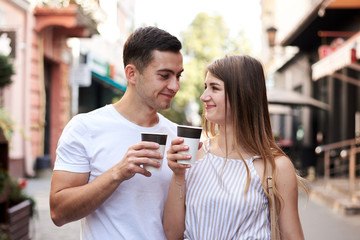 Romantic couple on date drinking coffee cappuccino in paper cups on street . Young blond woman, walking with brunette man with yummy coffee drink in summer in morning. Nutritious breakfast, snack.