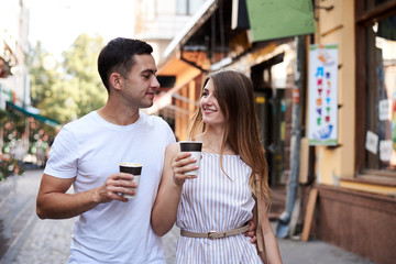 Romantic couple on date drinking coffee cappuccino in paper cups on street . Young blond woman, walking with brunette man with yummy coffee drink in summer in morning. Nutritious breakfast, snack.