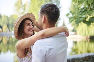 Fototapeta premium Young couple in love, hugging in the park in summer, smiling. Pretty blond girl in stripy overall and straw hat on romantic date with handsome brunette guy in white t-shirt. Sunday city walk.