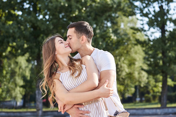 Handsome brunette guy, wearing white t-shirt, hugging and kissing pretty blond woman in stripy overall. Young couple in love, embracing in green park in summer. Romantic relationship. Valentines day.