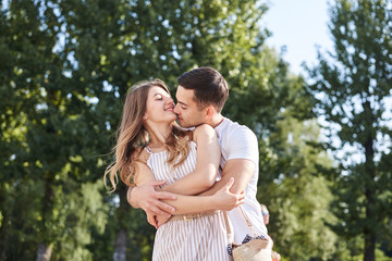Fototapeta premium Handsome brunette guy, wearing white t-shirt, hugging and kissing pretty blond woman in stripy overall. Young couple in love, embracing in green park in summer. Romantic relationship. Valentines day.