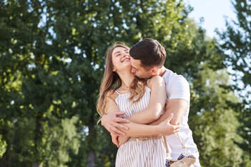 Handsome brunette guy, wearing white t-shirt, hugging and kissing pretty blond woman in stripy overall. Young couple in love, embracing in green park in summer. Romantic relationship. Valentines day.