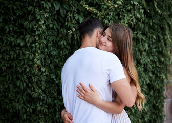 Young couple in love hugging near green bushes trees wall. Pretty blond woman, wearing stripy short overall and brunette man in white t-shirt and blue shorts on romantic date. Romantic relationship