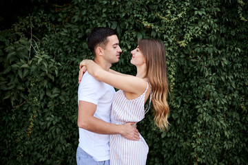 Young couple in love hugging near green bushes trees wall. Pretty blond woman, wearing stripy short overall and brunette man in white t-shirt and blue shorts on romantic date. Romantic relationship