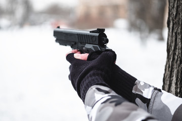 A military man in camouflage and gloves is aiming with a pistol next to a tree.
