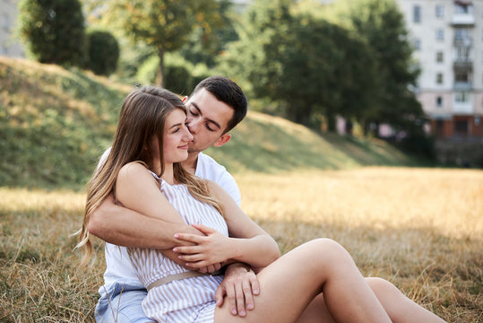 Young couple in love sitting on green yellow grass lawn hugging embracing kissing. Blond woman wearing stripy short overall and brunette man in white t-shirt blue shorts on romantic date. Relationship