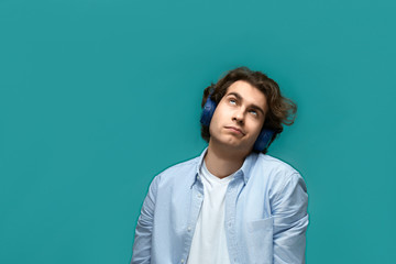 Fototapeta premium Portrait of a young beautiful man wearing white t-shirt and blue shirt in blue headphones rolled his eyes and looking up over blue background
