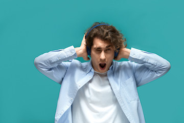 Portrait of a young beautiful man wearing white t-shirt and blue shirt in headphones covers ears with hands shouting from noise and looking on camera over blue background
