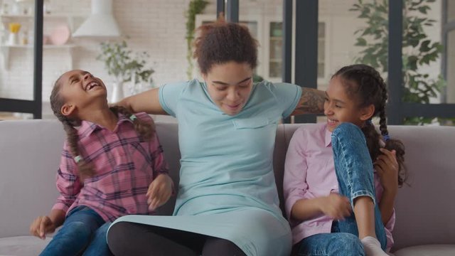 Joyful playful attractive african american mother and two cheerful preadolescent girls enjoying leisure together on couch in domestic room, tickling each other and having fun during weekend.