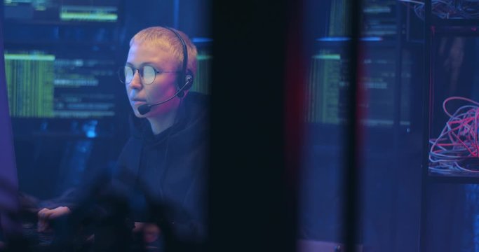 Caucasian woman from cyberpolice with headset and mic sitting in dark room with monitors at desk and working at computer at night. Female hacking online system. Stealing information concept.
