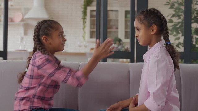 Carefree playful adorable elementary age african sisters sitting face to face on cozy couch, playing staring contest, gesturing , tickiling each other and trying to force to blink in domestic room.