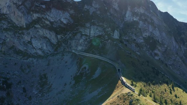 Follow aerial shot of unique switzerland railway train going uphill through tunels in mountains. Green grass with blue sky. Cableway lift for skiers at background