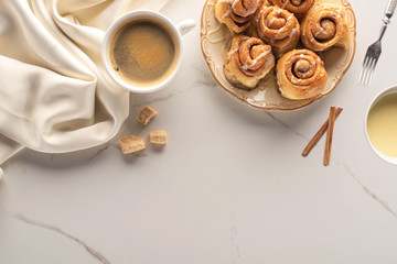 top view of fresh homemade cinnamon rolls on marble surface with cup of coffee, brown sugar, condensed milk, fork and silk cloth