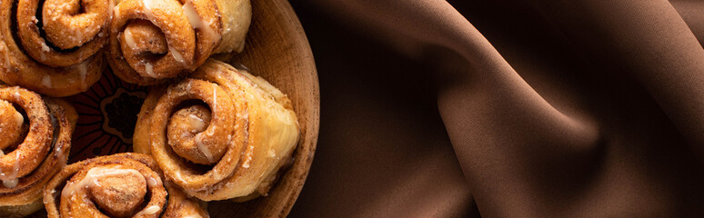 top view of fresh homemade cinnamon rolls on plate on silk brown cloth, panoramic shot