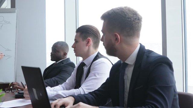 person stands by flipchart. A team of young businessmen in suits working and communicating together in an office. Corporate businessteam and manager in a meeting.