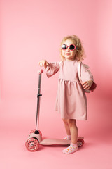 A girl in sunglasses and a pink dress with a backpack on her back rolls on a pink scooter.