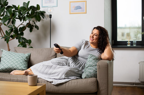people and leisure concept - happy young woman lying on sofa with remote control and watching tv at home