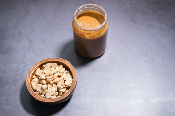 Peanut butter in a glass jar with some peanuts in a wooden bowl on concrete backgroud. Copy space.