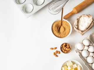 Set of various baking ingredients - flour, eggs, sugar, butter, nuts, kitchen utensils and cupcake baking dish on white background. Top view.