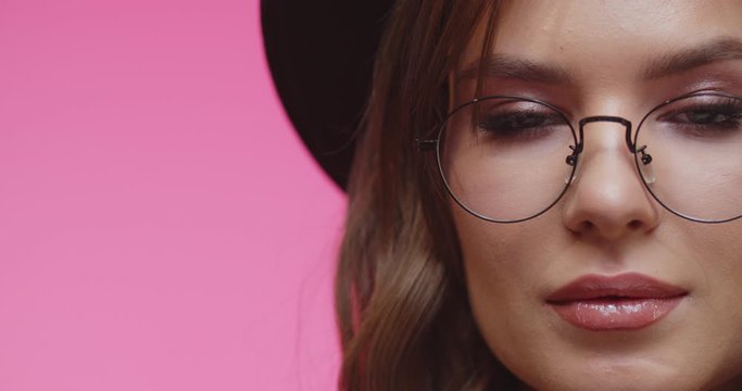 Model with blach hat and glasses looking at camera . Cheerful stylish brunette girl in hat looking in camera in studio with pink background. Female looking into camera in close up.