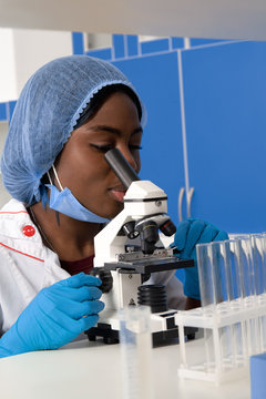 Young African Female Tech Or Scientist In Protective White Coat, Hat, Gloves And Mask Works With Microscope In Modern Laboratory Or Research Facility. Analyzing Patient Biopsy Samples, Diagnose Cancer