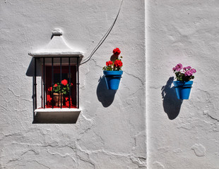 Fachada típica andaluza con ventana con geranios y macetas azules con flores colgadas en la pared