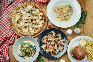 A variety of dishes for lunch - pizza, pasta carbonara, hamburger, stewed vegetables and salad with jamon. Top view, food flat lay. Dinning table