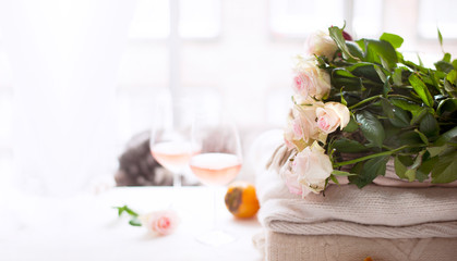 Fototapeta na wymiar Bouquet of roses natural light. Flowers by the window and two glasses of rose wine. Cozy and romantic atmosphere. Banner long format. Copy space. Selective focus shifted from the subject.