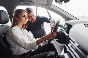 Woman testing car by help of male assistant indoors in the salon