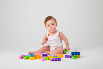 baby with a pacifier in his mouth in white clothes sitting playing with a multi-colored constructor on a white background, space for text