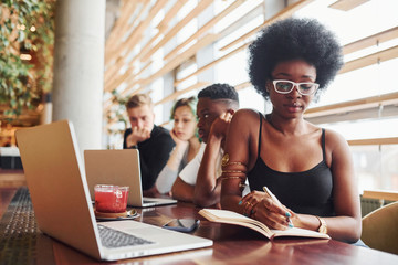 Black woman sitting in front of group of multi ethnic people with alternative girl with green hair is working together by the table indoors