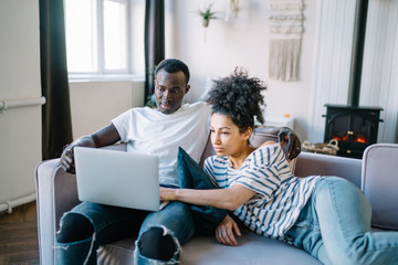 Multiracial couple comfortably sitting on sofa with credit card and looking at laptop