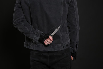 Man with knife behind his back on black background, closeup. Dangerous criminal