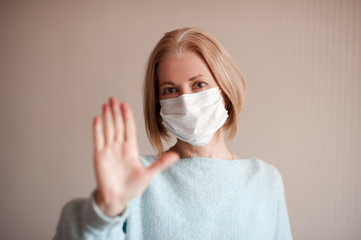 Adult woman 45-50 year old wearing medical mask making stop sign with hand posing in room closeup. Coronavirus concept. Healthy lifestyle. Looking at camera.