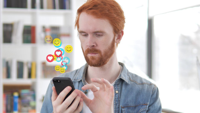Redhead Beard Designer Using Smartphone, Flying Emojis, Smileys and Likes