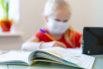 Distance learning online education. Blurred sickness schoolgirl in medical mask studying at home with digital tablet in hand and doing school homework. Training books and notebooks on table