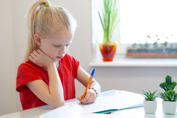 Serios blonde schoolgirl sitting at home and doing school homework, writing in notebook. Training...