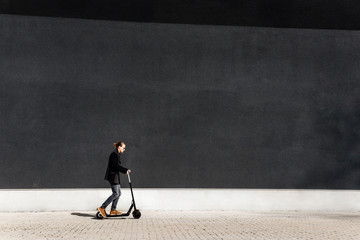 A young man rides an electric scooter along a large black wall.